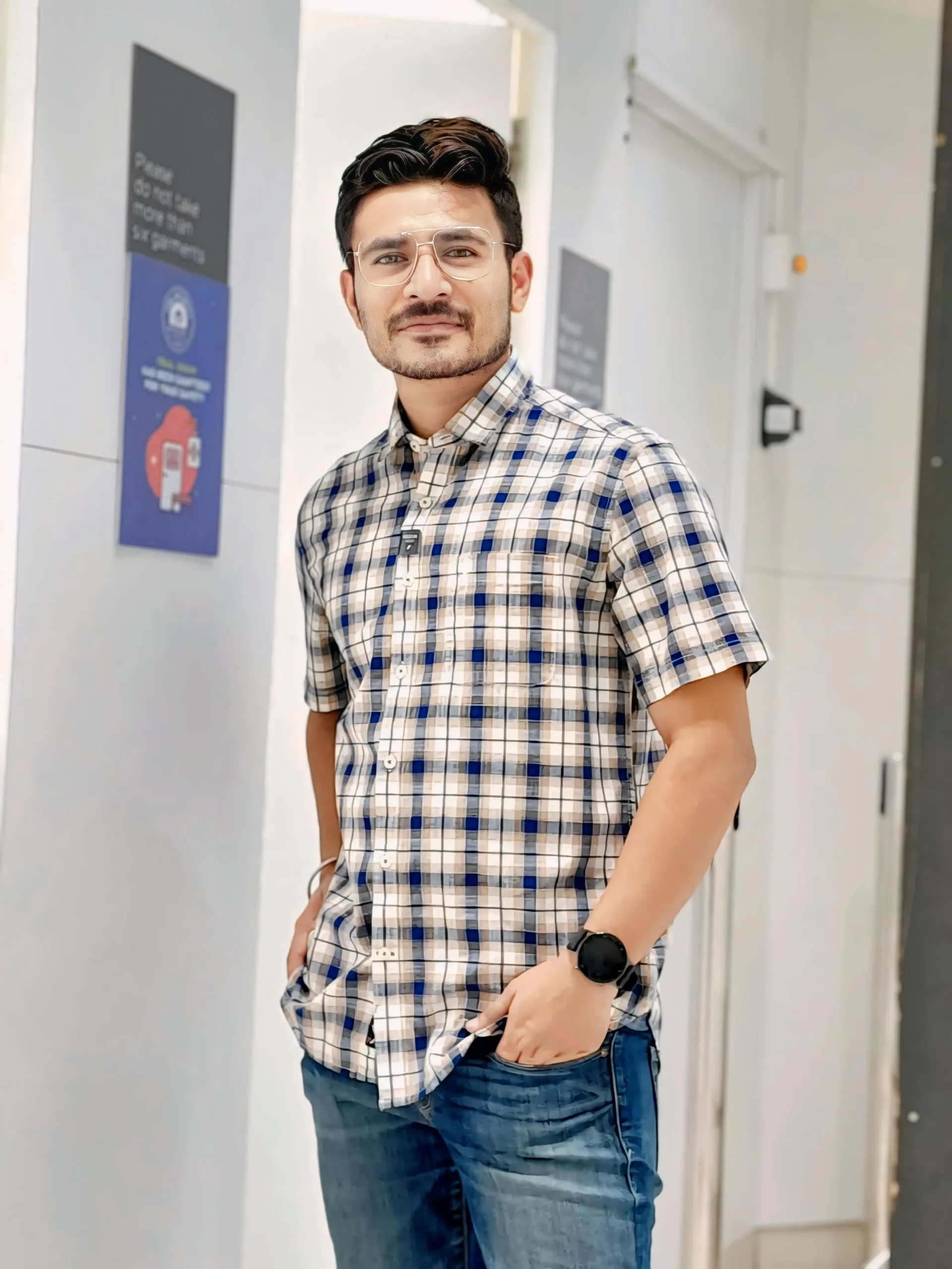 Rohit Upadhya Founder of Digitophile wearing a plaid shirt and glasses stands casually with one hand in his pocket, looking at the camera in a modern indoor setting.